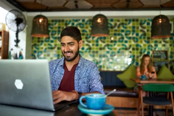 Handsome millennial indian man working in trendy coworking space cafe on laptop drinking organic fair-trade cappuccino
